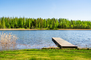 Lake and dock in Finland. Wood pier. Finnish nature landscape. Beautiful summer day. Water and blue sky. Forest in the background. Grass beach in vacation cottage.