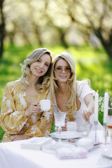 There are two women happily sitting at a table while holding cups of tea