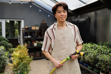 Smiling asian florist holding watering hose and looking at camera in greenhouse