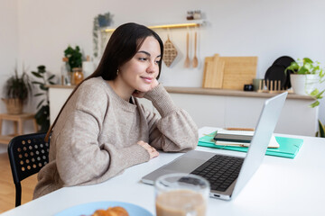 Young student woman studying online, listening teacher on laptop video call from kitchen at home. Education concept