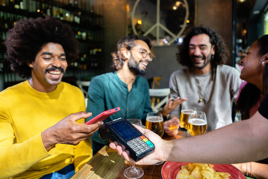 Happy young African American man using nfc technology on mobile phone to pay for drinks at restaurant, enjoying happy hour with diverse friends