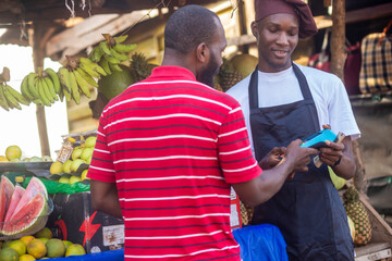 Person paying for fruits with a credit card while african fruit seller holding an electronic POS...