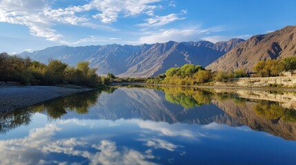 Fototapeta premium Serene river reflecting a tranquil mountain landscape. Tranquil waters mirror the majestic peaks and clouds. Autumn colors paint the valley's edges