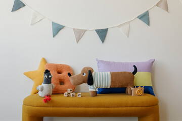 Soft toys and graphic pillows arranged on a mustard bench under a pastel bunting — playful setup in a cozy children room.