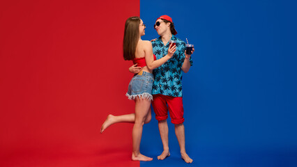 Young happy man in summer clothes hugging his beautiful girlfriend, couple enjoying positive atmosphere against red and blue background. Concept of human emotions, relationship, lifestyle