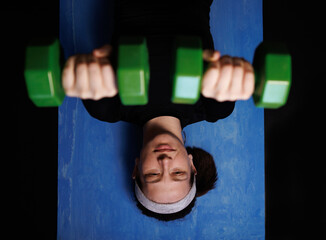 young woman with dumbbells in her hands performs home workout. the girl lies on a blue sports mat...