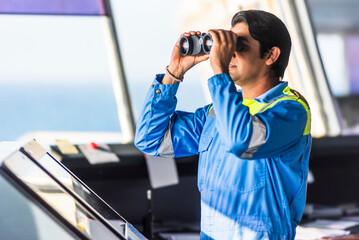 Chief officer in light blue uniform stands on ship's bridge, observing through binoculars by the...
