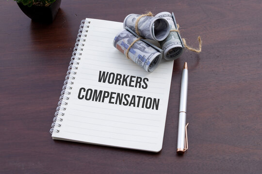  A notebook labeled "Workers Compensation" accompanied by cash, symbolizing financial planning and legal considerations, rests on a rustic wooden table.