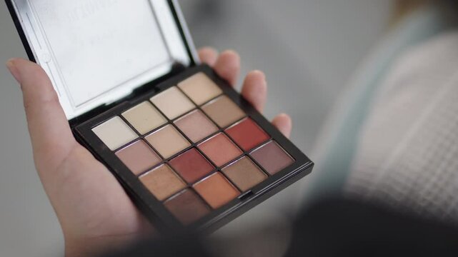 Close-up of hands applying makeup using a brush and a warm-toned eyeshadow palette.