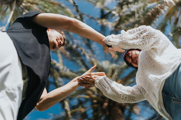 A low-angle view of a couple holding hands, smiling and enjoying a sunny day near palm trees....