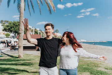 A cheerful couple embracing and enjoying their time together at a tropical beach under clear blue skies, with palm trees and the sea in the background.