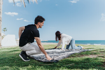 A man and woman arranging a blanket on grassy terrain near a sunny beach, enjoying the outdoors and...