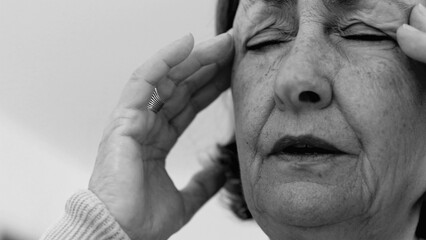 Elderly woman in close-up, eyes shut, with both hands pressing against her temples, deep wrinkles...
