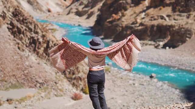 Young Indian woman walk near Blue Tsarap river at Phugtal monastery trek in Zanskar, Ladakh, India. Tourist woman raising hands against Chenab river flowing in valley. Travel and holidays concept