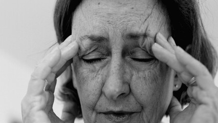 Elderly woman in close-up, eyes shut, with both hands pressing against her temples, deep wrinkles...
