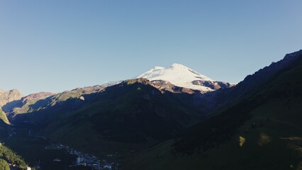 Fototapeta premium The snow-capped peak of Elbrus rises above the mountain valley in the rays of the rising sun. Mountains with a town in the lowland aerial view from a quadcopter