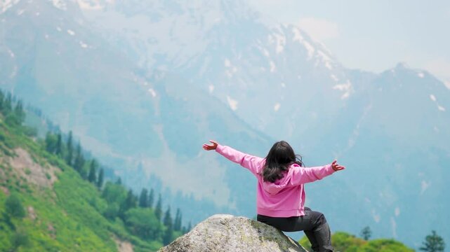 Indian girl wearing pink hoodie sitting against mountains. Happy people in nature. Teenager girl sitting outside, arms raised. Dreaming, freedom and traveling concept. Female mental health wellness