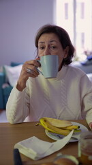 Older woman sipping from a cup of coffee while sitting at a table. Morning routine captured in a...