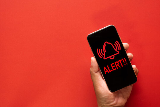 Hand holding mobile phone displaying alert notification, red bell icon with "ALERT!!" message, phone warning, emergency notification, technology, communication, urgent message concept.