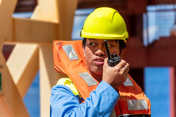 Merchant ship officer in blue overalls and yellow helmet, wearing lifejacket,  explains lifeboat...