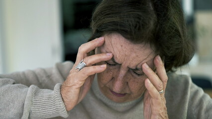 Senior woman in visible distress, holding head with eyes closed, fighting severe headache. Facial...