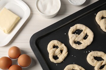 Baking tray with raw pretzels and ingredients on white wooden table, flat lay