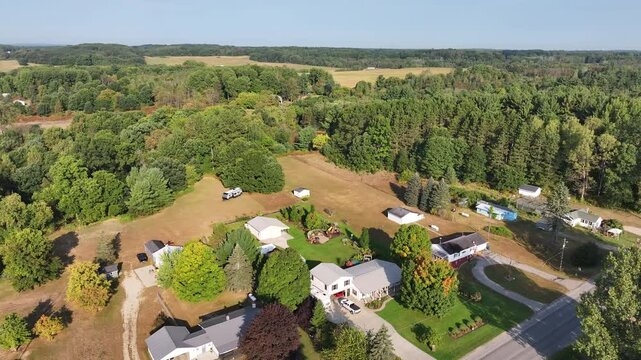 Aerial view above a two floor rural home with a truck parked outside and a fire pit, playground, shed, pole barn, and garden surrounded by woods, forest, a major road and farmland under a blue sky.