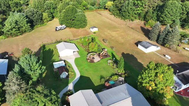 Aerial view above a two floor rural home with a truck and camper parked outside and a fire pit, playground, shed, pole barn, and garden surrounded by woods, forest, and farmland underneath a blue sky.