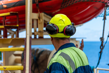 Marine engineer, merchant ship officer in blue overalls and yellow helmet explains lifeboat...