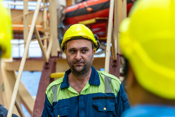 Marine engineer, merchant ship officer in blue overalls and yellow helmet explains lifeboat...