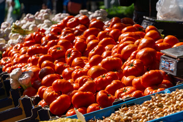 stand at the market with tomatoes and other vegetables. Fresh, red tomatoes are visible in the foreground, and garlic and onions in the background