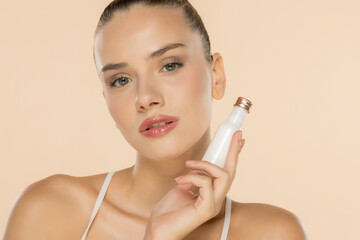 Beautiful woman with natural makeup holding white cosmetic bottle, showcasing skincare product against beige background.