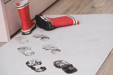 Red rubber boots stand on a welcome mat, marking a clear trail of muddy footprints across its...