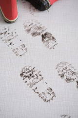 Red rubber boots stand on a welcome mat, marking a clear trail of muddy footprints across its...