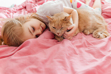 Child plays and cuddles with a friendly orange cat on a pink bedspread during the day