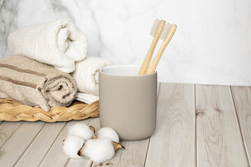 Eco-friendly wooden toothbrushes in a glass on a light wooden table with soft towels and cotton. Natural bathroom accessories for sustainable lifestyle and zero waste concepts.