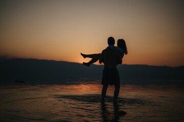 A silhouetted couple enjoying a romantic moment by the water during sunset. The peaceful scenery...