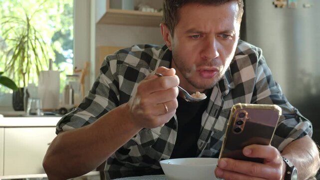 Man in plaid shirt eating oatmeal porridge from white bowl while using smartphone. Male looking at phone during breakfast. Concept of social media addiction, distraction, and digital dependency