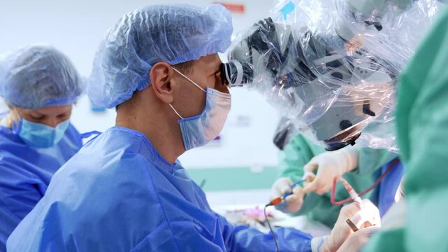 Concentrated male surgeon looks at microscope. Assistant cleans the tool device at backdrop and doctor gives him a used instrument.