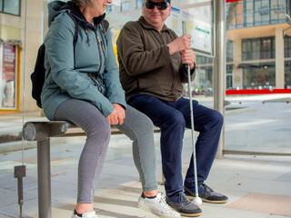 Blind man with a white cane speaks with an unrecognizable woman at the bus stop.