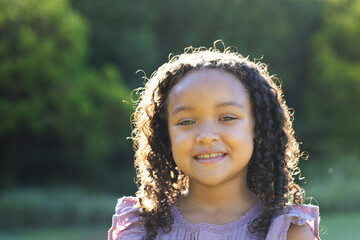 Smiling child girl standing outdoors in sunlit grassy area, with dark curly hair halo