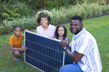Smiling diverse family kneeling on lawn at backyard garden, with large solar panel for energy