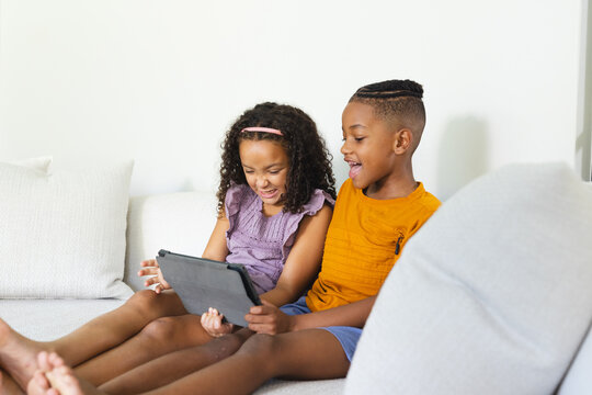 Smiling Diverse siblings using tablet device on light-colored sofa in living room, with cushions