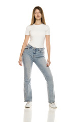Obraz premium Young woman with long straight brown hair wearing white fitted t-shirt and light blue bootcut jeans with white sneakers, against white background, full body