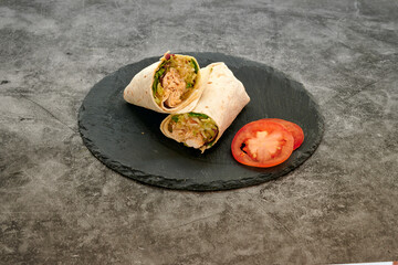 A wrap with a tomato on top of it. The wrap is on a black plate. The plate is on a gray surface