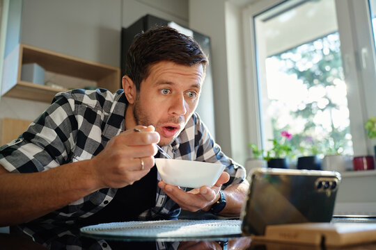 Surprised man in checkered shirt has breakfast at kitchen table, eats porridge from bowl, watching online content on his smartphone. Concept of screen addiction and distraction