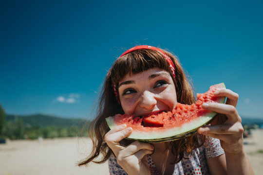 A cheerful young woman bites into a watermelon slice on a bright summer day, capturing the joy of nature and outdoor fun near a scenic beach and lake environment.