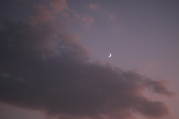 A serene twilight sky with soft, purple-tinged clouds and a delicate crescent moon glowing above....