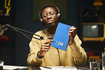 African American man in headphones sitting at table with microphone and discussing book during...