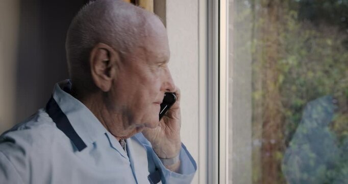 Senior man, window and phone call at window with fear, talking about emergency, intruder or panic while upset, suspicious and worried. Old person with in communication waiting for security help